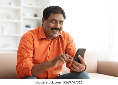 Closeup of happy handsome mature indian man wearing casual outfit sitting on couch at home, using phone and smiling, chatting with beloved wife or kids, checking news. Nice online offer, deal - Powered by Shutterstock