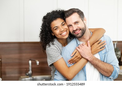 Close-up of happy interracial couple posing over blurred kitchen background, happy owners of new flat smiling and looking at the camera, young African American woman hugging handsome man from behind - Shutterstock ID 1912555489