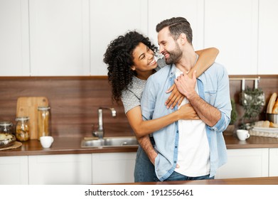 Close-up of a happy interracial couple standing in the kitchen, happy romantic owners of a new flat smiling and looking at each other, young African American woman hugging handsome man from behind - Shutterstock ID 1912556461