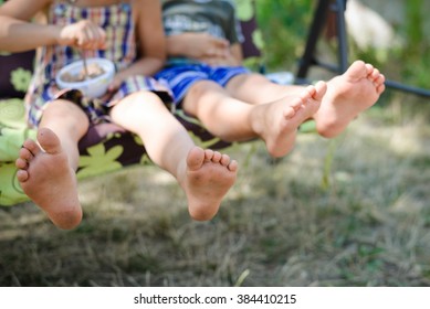 Closeup Of Happy Kids Sitting And Swing On Sunny Countryside Background. Picture Of Two Children Legs Barefoot. 