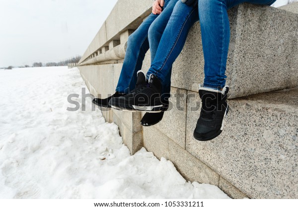 Close-up of a happy and loving couple foot\
traveler sitting on the granite waterfront. A pair in winter shoes\
and warm sweaters and jeans, dangling with their feet, dangling in\
the winter landscape
