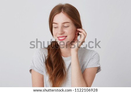 Closeup of happy playful young woman with long red hair and freckles touching her ear and smiling isolated over white background Looking to the side