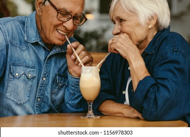 Closeup Of Happy Senior Couple In Love At Cafe Drinking Cold Coffee From Same Glass. Old Man And Woman Enjoying A Cold Coffee On Table.