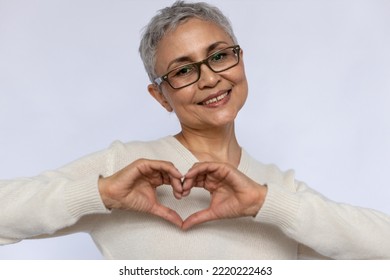 Close-up Of Happy Senior Woman Showing Gratitude Symbol. Mature Caucasian Woman Wearing Eyeglasses And White Jumper Looking At Camera And Smiling Over White Background. Gratitude, Love, Health Symbol