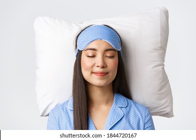 Close-up of happy smiling beautiful asian girl in sleeping mask and blue pajamas, lying in bed on pillow with pleased expression, dreaming about something satisfying, white background - Powered by Shutterstock