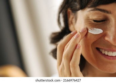Close-up happy young Caucasian woman smiling with her eyes closed applying cream on her face. Brunette moisturizes her skin with cosmetics. Home spa care concept - Shutterstock ID 2166976427