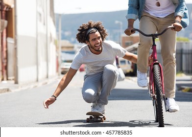 Closeup Of A Happy Young Man Skateboarding With Help Of Friend Bicycle. Smiling Guys Having Fun With Skate Board And Cycle. African Man Riding A Bike While His Friend Attacking With Skateboard.