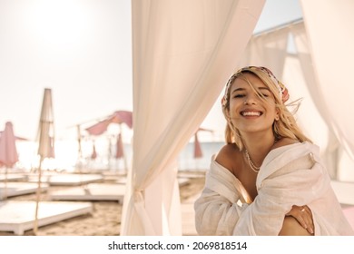 Close-up of happy young woman smiles broadly on blurred beach background lined with sun loungers with umbrellas. She is wearing loose white shirt and headscarf on her head.