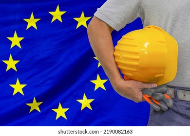 Close-up Of Hard Hat Holding By Construction Worker On EU Flag Background. Hand Of Worker With Yellow Hard Hat And Gloves. Concept Of Industry, Construction And Industrial Workers In European Union