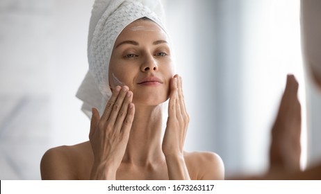 Closeup head shot pleasant beautiful woman applying moisturizing creme on face after shower. Smiling young pretty lady wrapped in towel smoothing perfecting skin, daily morning routine concept.