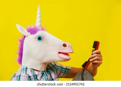 closeup of the head of a young man wearing a unicorn mask, talking on a red landline telephone, against a yellow background - Shutterstock ID 2124901742
