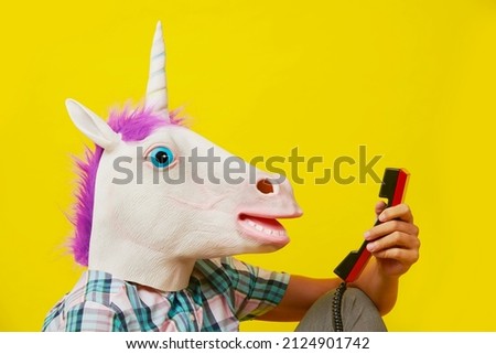 closeup of the head of a young man wearing a unicorn mask, talking on a red landline telephone, against a yellow background