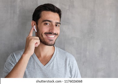 Closeup headshot of handsome European Caucasian guy isolated on gray background wearing wireless headphones and listening to media files or talking to someone, touching his ear with happy smile