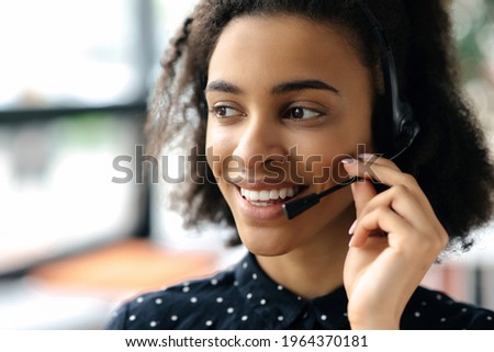 Close-up headshot of pleasant charming young african american curly haired woman in headset and stylish shirt, female call center worker or support operator, looking to the side, smiling friendly