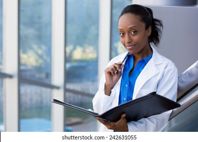 Closeup Headshot Portrait Of Friendly, Smiling Confident Female Doctor, Healthcare Professional With Labcoat, Holding Pen To Face And Holding Notebook Pad. Isolated Hospital Clinic Background.