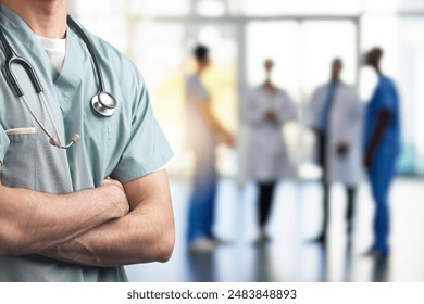 A close-up of a healthcare professional in scrubs with arms crossed and a stethoscope around their neck, with a blurred background of medical staff in a hospital or clinic setting. - Powered by Shutterstock