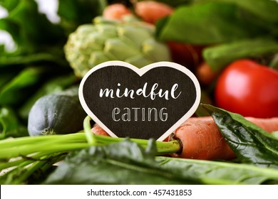 closeup of a heart-shaped chalkboard with the text mindful eating placed on a pile of some different raw vegetables, such as cucumbers, tomatoes, carrots, artichokes, green peppers and zucchinis