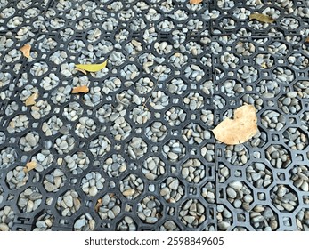 Close-up of a hexagonal plastic grid filled with pebbles in a garden. There are dry leaves scattered around.