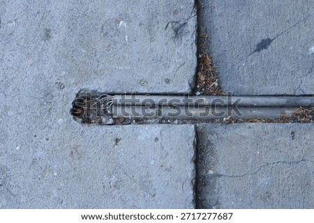 Close-up high angle view of two weathered plastic conduits embedded in a gap between cracked and uneven concrete pavement slabs. The conduits run horizontally across the frame, partially filled with dirt and debris. On the left side, a section of one conduit is damaged, revealing a bundle of insulated wires. The overall texture is rough and industrial, highlighting the exposed infrastructure beneath the surface.