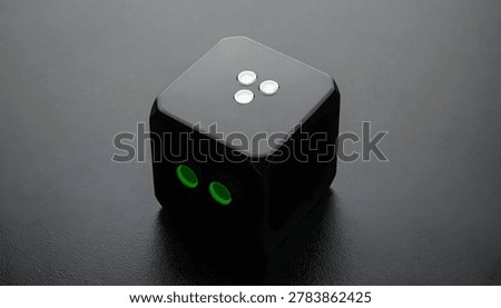 A close-up, high-angle shot of a black cube with glowing green circular lights on one side and three white dots on the top, set against a dark, textured background.