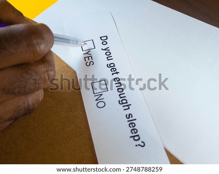 A close-up, high-angle shot captures a person’s hand poised to mark a paper survey. The individual, with a dark complexion, holds a clear ballpoint pen over a white slip of paper resting on a wooden surface. The printed text asks, 