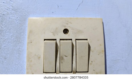 A close-up, high-angle shot captures a weathered, off-white triple light switch plate mounted against a textured, light-colored wall, revealing signs of age and neglect through the layer of dust and grime accumulation on its surface, while the single centered screw stands out as a focal point between the switches, emphasizing the mundane, utilitarian nature of this everyday household object.
