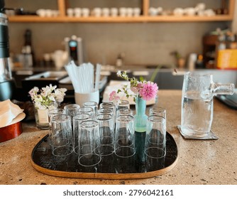 A close-up, high-angle shot of multiple clear empty water glasses neatly arranged on a wooden-rimmed black tray in a cozy cafe, accompanied by small vases with fresh flowers, creating a calm and minimalist atmosphere.