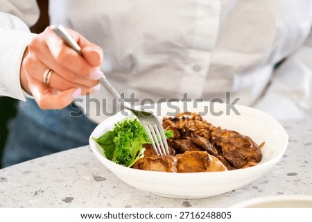 A close-up, high-angle shot of a person using a silver fork to eat a healthy meal of chicken or meat with rice and green lettuce in a white bowl