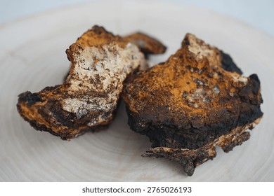 A close-up, high-angle shot of two large chunks of wild Chaga mushroom (Inonotus obliquus) resting on a light-colored, textured wooden surface. The image highlights the distinctive appearance of the medicinal fungus, featuring a dark, almost black, charcoal-like outer crust and a rich, golden-brown interior. The pieces show a rugged, porous texture with hints of lighter woody fibers where they were detached from the host birch tree. The lighting is soft and natural, emphasizing the organic details and earthy tones of the mushrooms. This photograph is ideal for content related to herbal medicine, natural superfoods, traditional healing, and forest foraging.