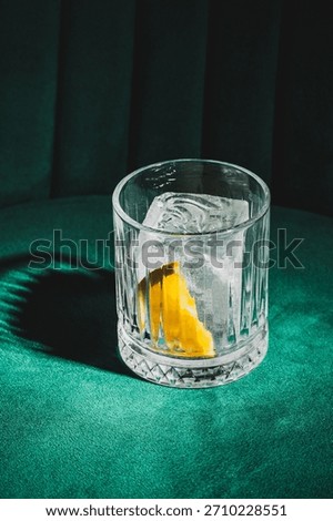 A close-up, high-angle studio shot of a clear old-fashioned cocktail or spirit on the rocks with a lemon twist, served in a textured crystal tumbler on a deep green velvet surface