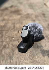 A close-up, high-angle studio shot showcasing a compact, modern wireless lavalier microphone designed for professional content creation. The device features a sleek, matte black finish with a prominent power button, integrated with a plush, grey faux-fur windscreen to effectively minimize wind noise during outdoor recordings. The microphone is attached to a sturdy, spring-loaded clip, allowing for easy and secure attachment to clothing. The unit rests on a rustic, weathered wooden surface, which creates a striking contrast between the modern, high-tech audio equipment and the natural, textured backdrop. The lighting is bright and directional, highlighting the intricate details of the fur texture and the solid construction of the device, making it an ideal visual asset for tutorials, tech reviews, blogs, and marketing materials focused on mobile videography, podcasting, and digital content production gear.