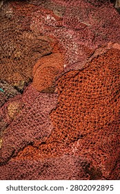 A close-up, high-angle view captures a chaotic pile of various fishing nets. The nets display a range of sun-faded reds, oranges, and deep browns, highlighting their intricate woven patterns and rugged texture.