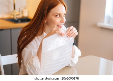 Close-up high-angle view of happy young woman playing pops bubble wrap to calm herself sitting at table in kitchen. Smiling pretty redhead female popping plastic bubblewrap. Concept of mental health.