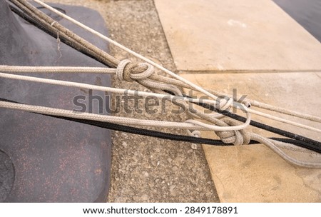 Close-up high-angle view of multiple thin white and black nautical ropes secured with knots around a heavy harbor post on a concrete pier. Ideal for maritime industry articles, vessel rigging topics, sailing techniques, port security, and marine engineering tutorials.