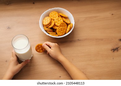Close-up high-angle view of unrecognizable little child girl taking cookie from plate sitting at table with glass of milk. Cropped shot of kid making snack during online distance learning at home.