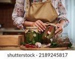 Close-up of homemade pickled canned cucumbers and chili peppers in sterilized glass jars in the hands of a young woman housewife. Fresh fragrant ingredients and culinary herbs on a kitchen table