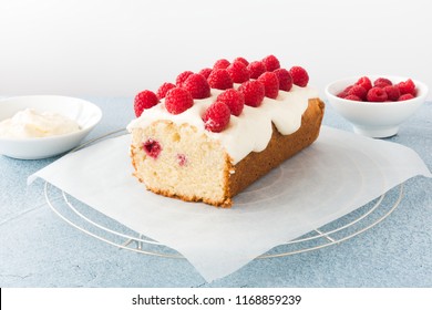 Closeup of a homemade raspberry loaf cake on parchment paper and cooling rack. Bowls filled with fresh raspberries and frosting in the background.