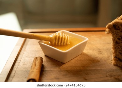 A close-up of a honey dipper resting in a small dish of honey on a wooden cutting board. The scene includes a cinnamon stick and a partially visible loaf of cranberry bread in the background. - Powered by Shutterstock