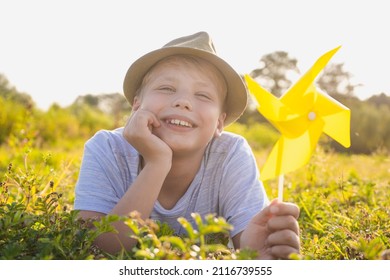 Closeup Horizontal Color Photography Of Happy Child. Portrait Of Smiling White Kid Relaxing Happily In Sunny Sunset Meadow Outside. Boy Holding Yellow Plastic Pinwheel Toy In Hand