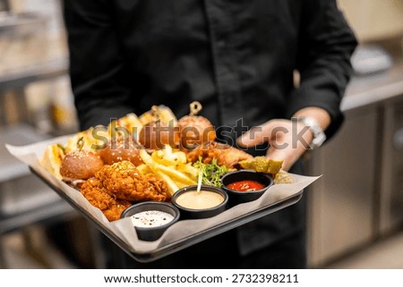 A close-up, horizontal, eye-level shot of a server holding a black tray laden with an appetizer platter of mini burgers, fried chicken, french fries, and dipping sauces.