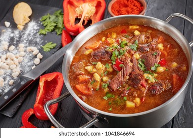 Close-up Of  Hot Beef Hungarian Goulash  Or Bograch Soup With Paprika, Small Egg Pasta, Vegetables And Spices In A Pot. Ingredients And Soup Ladle On Table, Classic Recipe, View From Above, Close-up