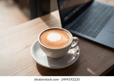 Close-up of Hot coffee latte with latte art milk foam in cup mug and laptop computer on wood desk office desk in coffee shop at the cafe,during business work concept