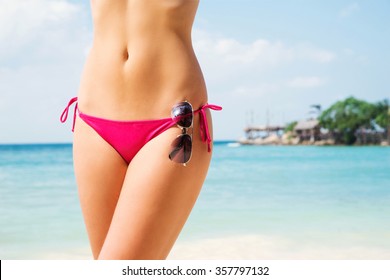 Close-up of hot and seductive lady with perfect figure in magenta panties posing over sea background with sunglasses.