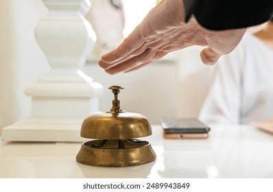 Close-up of hotel reception desk featuring antique brass service bell. Guest's hand reaching to ring for assistance, symbolizing luxury hospitality and customer service.