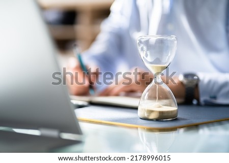 Close-up Of Hourglass In Front Of Businessperson's Hand Calculating Invoice
