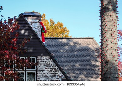 Closeup Of House With Christms Lights On Roof And Tree And Deflated Blow-up Santa Hanging From Chimney