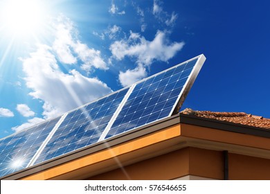 Close-up Of A House Roof With A Solar Panels On Top, On A Blue Sky With Clouds And Sun Rays