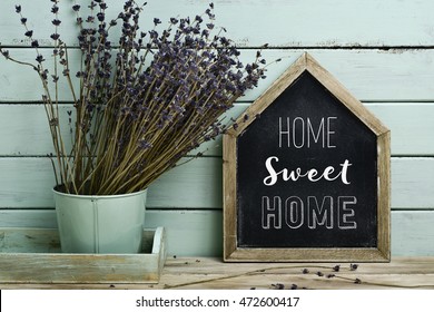 closeup of a house-shaped chalkboard with the text home sweet home written in it and a bunch of lavender flowers in a flower pot, against a rustic pale blue background