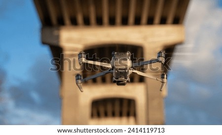 Close-up of a hovering drone with a camera, captured in clear focus against a blurry bridge backdrop during sunrise.