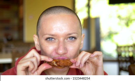 Close-up. Hungry Bald Woman In A Restaurant Eating Delicious Chicken Wings. Looking At The Camera.